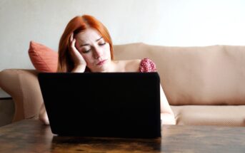 A woman with red hair wearing a red polka dot dress rests her head on her hand, looking tired or stressed, while sitting on a beige couch in front of an open laptop on a wooden table. A coral-colored pillow is in the background.