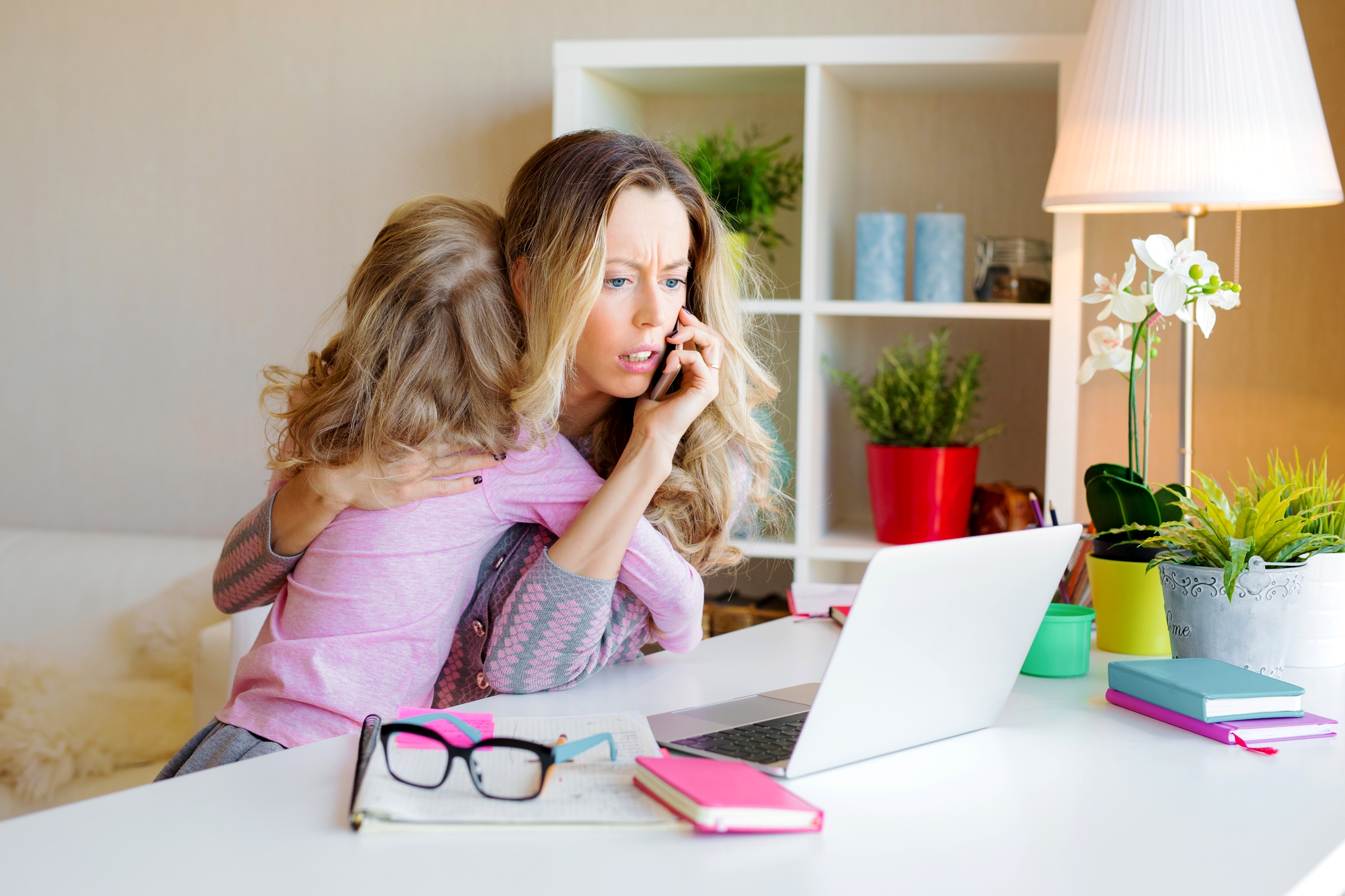 A woman sits at a desk, talking on the phone with a concerned expression, while holding a child in her lap. The desk has a laptop, notebook, glasses, and various plants. The room is brightly lit with a table lamp and decorated with plants and colorful items.