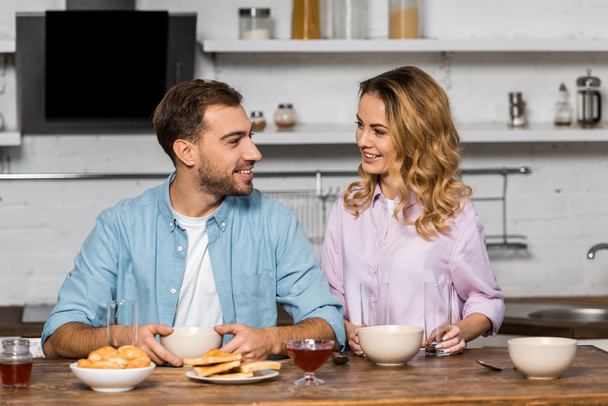 A man and a woman are sitting at a kitchen counter, smiling at each other. They are enjoying a meal, with bowls, plates of toast, and jars of jam in front of them. The kitchen has a modern design with white walls and shelves in the background.