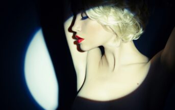 A woman with short blonde hair and red lipstick poses with her arms raised above her head. She is illuminated by a focused, blue-tinted spotlight, creating dramatic shadows against a dark background. Her eyes are closed, and she conveys a feeling of poise and elegance.