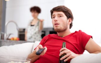 A man in a red shirt sits on a couch holding a TV remote and a beer bottle, looking intently at the television. A woman in the background, standing in the kitchen, appears to be talking or gesturing towards the man. A bowl of popcorn is in the foreground.
