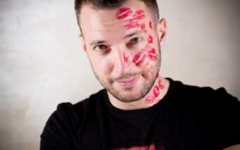 A man with short hair and a beard is smiling at the camera. His face and neck are covered in numerous red lipstick kiss marks. He is wearing a black t-shirt and has a playful expression. The background is a plain, light-colored wall.