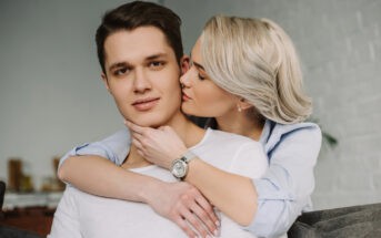 A woman with short blonde hair and a blue shirt is embracing a man with short brown hair and a white shirt from behind. She is gently kissing his cheek while they both sit on a gray couch in a cozy, well-lit living room.