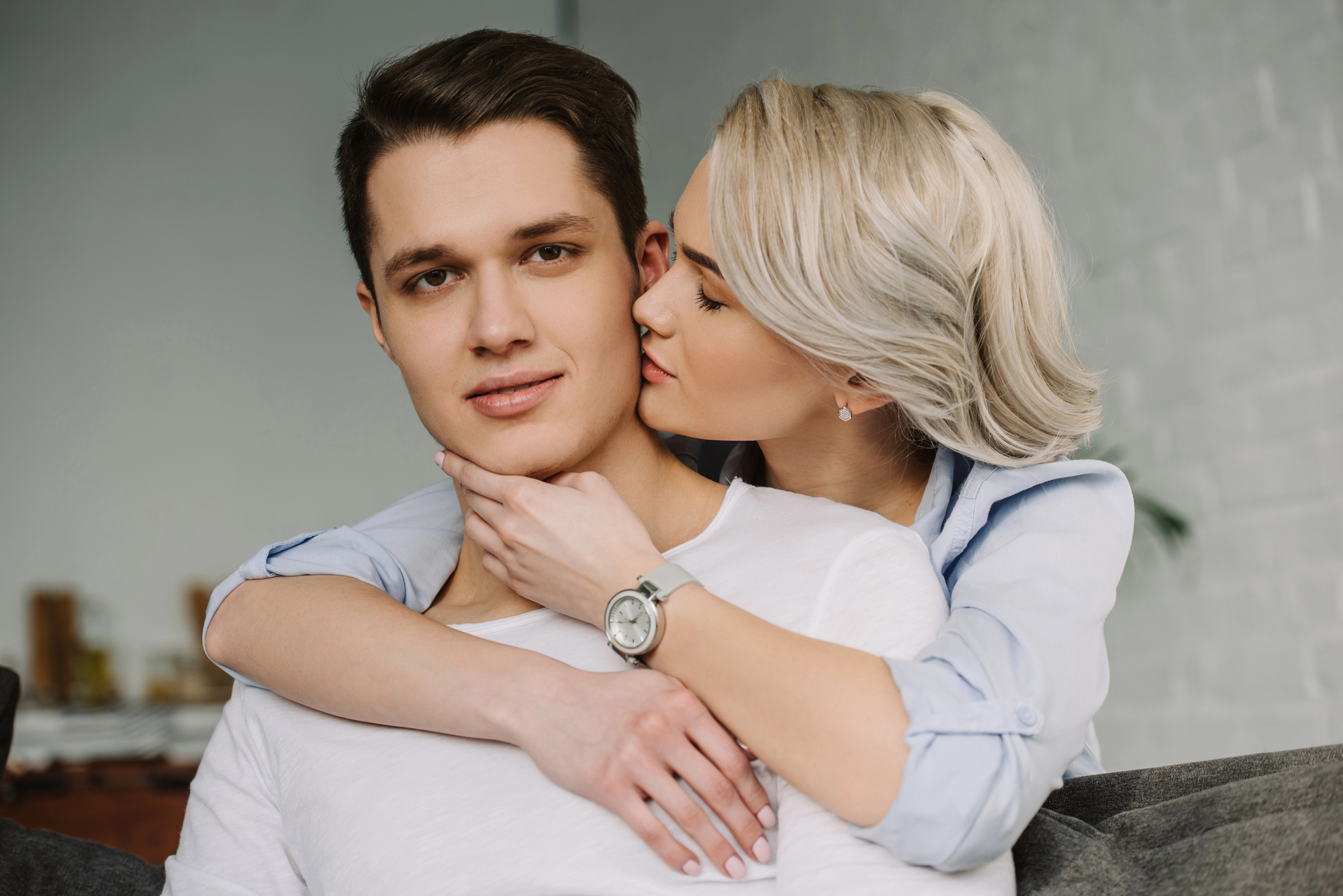 A woman with short blonde hair and a blue shirt is embracing a man with short brown hair and a white shirt from behind. She is gently kissing his cheek while they both sit on a gray couch in a cozy, well-lit living room.