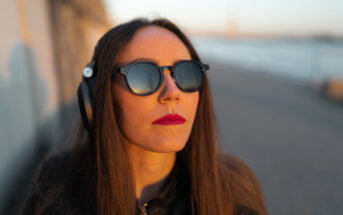 A person with long brown hair is wearing round sunglasses and headphones, looking up towards the sky. They have red lipstick and are standing outside, with a blurred background of a pathway and a river.