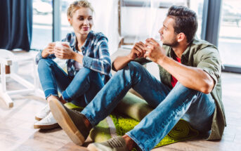 A man and a woman are sitting on the floor facing each other, casually talking and holding cups of coffee. They both appear relaxed and comfortable, dressed in casual clothing with jeans and shirts. Natural light streams in from the window behind them.