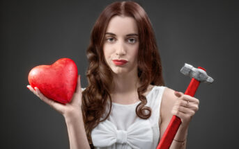 A woman with long, wavy brown hair is holding a red heart in her right hand and a hammer in her left hand. She is dressed in a white blouse with a large bow, and her facial expression is neutral, set against a dark grey background.