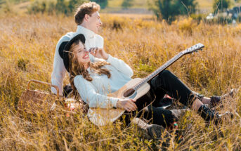 Two people are sitting in a sunlit field with tall grass. One person, wearing a hat and a white sweater, is holding a guitar and smiling. The other, also in a white sweater, is gazing into the distance. A picnic basket lies beside them. The atmosphere is relaxed and joyful.