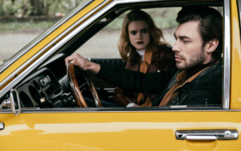 A man with dark hair and a beard is sitting in the driver’s seat of a yellow vintage car, holding the steering wheel and looking forward. A blonde woman in the passenger seat looks concerned as she watches the man. Both are wearing brown and black jackets.