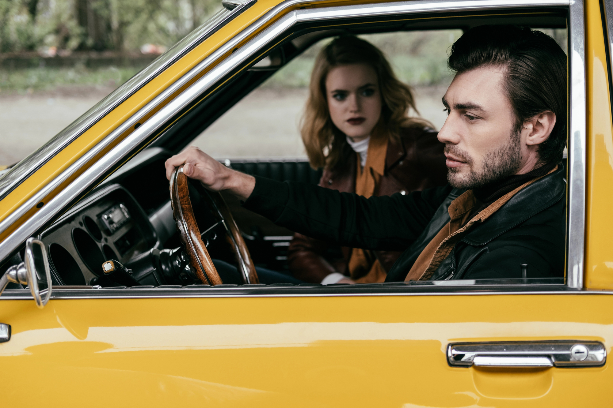 A man with dark hair and a beard is sitting in the driver’s seat of a yellow vintage car, holding the steering wheel and looking forward. A blonde woman in the passenger seat looks concerned as she watches the man. Both are wearing brown and black jackets.