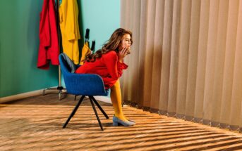 A woman in a red dress and yellow tights sits thoughtfully on a blue chair, gazing through vertical blinds. Sunlight streams in, casting striped shadows across the floor and her legs. The background features a teal wall with brightly colored coats and an umbrella.