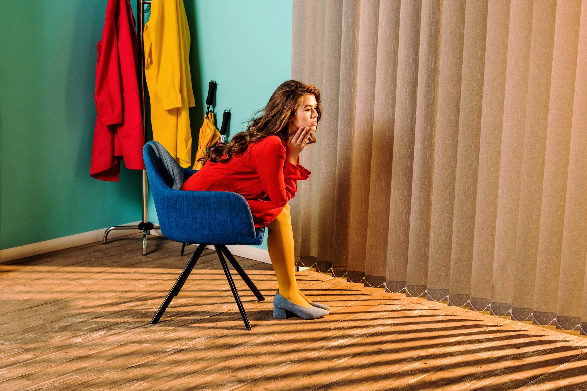 A woman in a red dress and yellow tights sits thoughtfully on a blue chair, gazing through vertical blinds. Sunlight streams in, casting striped shadows across the floor and her legs. The background features a teal wall with brightly colored coats and an umbrella.