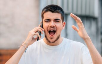 A young man with short hair and a beard is holding a smartphone to his ear with one hand, looking angry and shouting. His other hand is raised in frustration. He is wearing a white t-shirt and several bracelets on his wrist. The background is blurred.