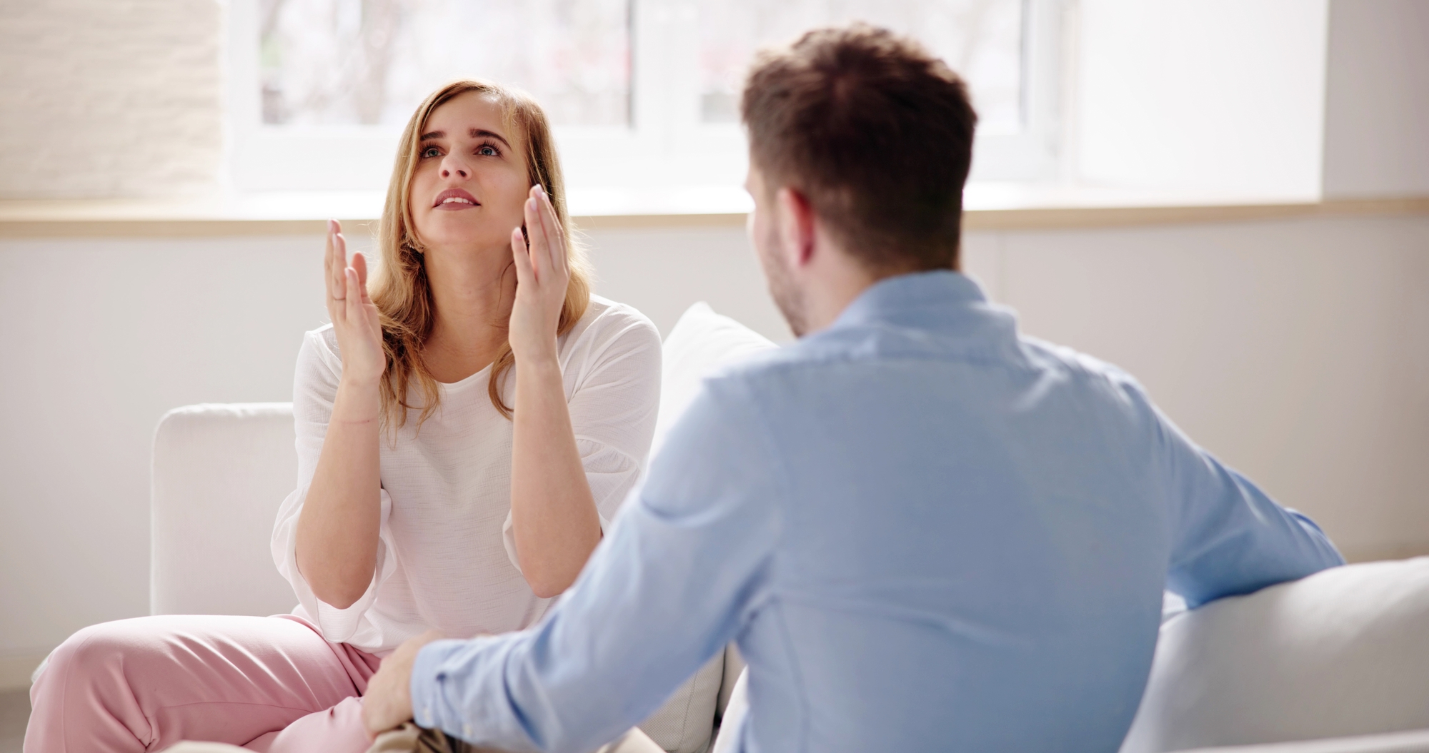 A woman with long brown hair, wearing a white top and pink pants, sits on a couch gesturing with her hands up, looking distressed. She is talking to a man with short hair wearing a light blue shirt and beige pants, who is facing her with his back to the camera.