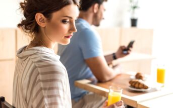 A woman with brown hair wearing a striped shirt holds a glass of orange juice while looking to the side, sitting at a table. In the background, a man with dark hair and a blue shirt looks at his phone. Croissants and orange juice are on the table.