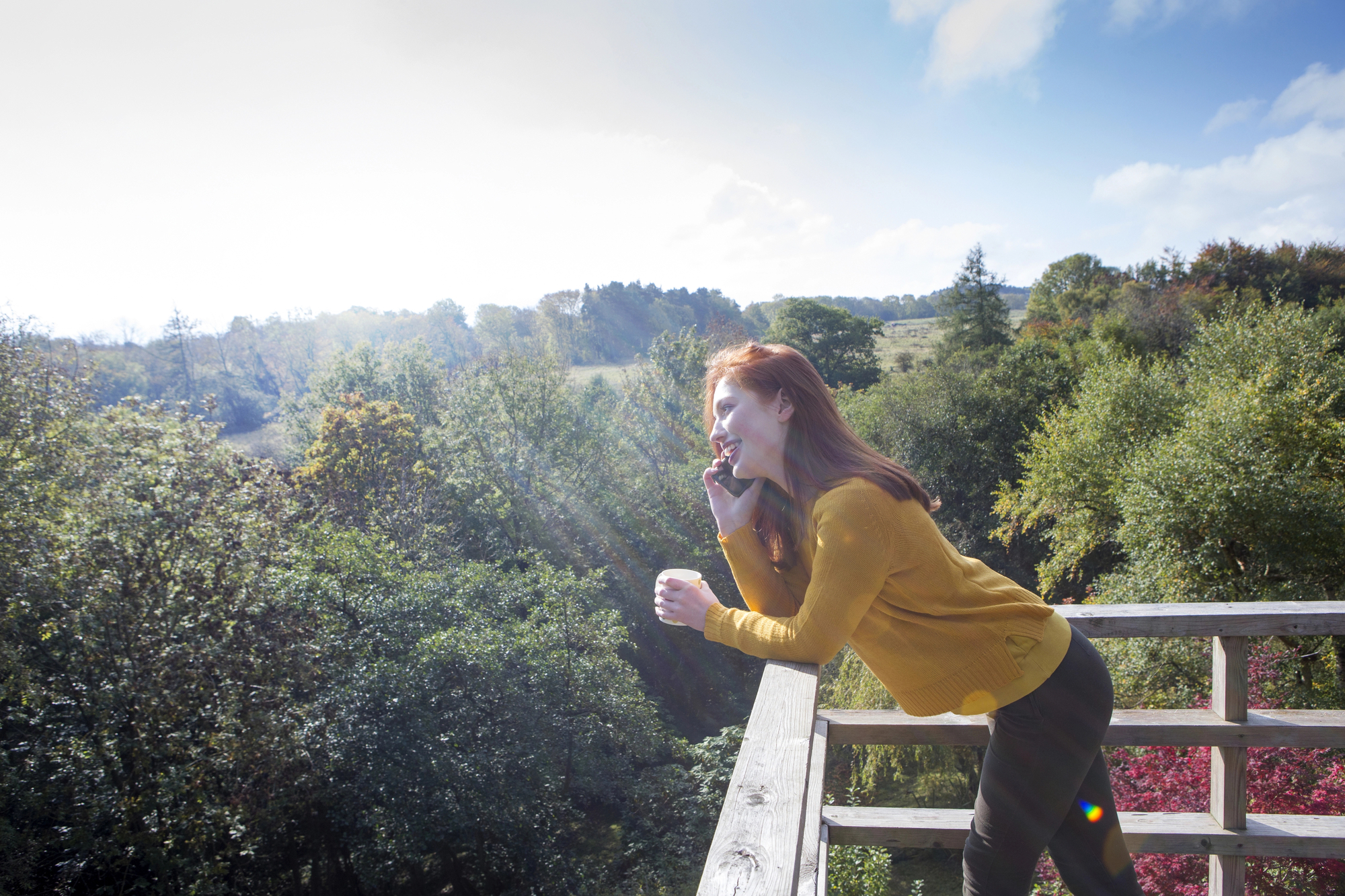 A woman with red hair leans over a wooden railing on a balcony, smiling while talking on her phone. She is wearing a mustard yellow sweater. The background shows a scenic view of lush green trees under a clear, sunny sky.