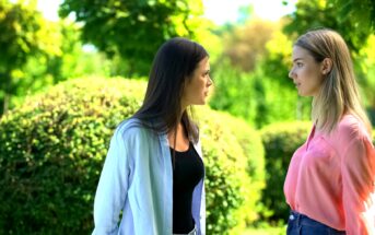 Two women are standing face-to-face in a park, engaged in a serious conversation. The woman on the left has long dark hair, a black top, and a light jacket. The woman on the right has blonde hair and is wearing a pink shirt and blue jeans. Greenery surrounds them.