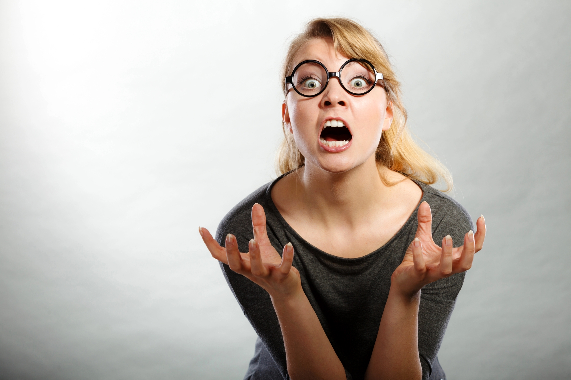 A person with blonde hair wearing glasses and a gray shirt has an expression of shock or surprise. Their mouth is open wide, and their hands are raised in front of them with fingers spread apart. The background is plain and light-colored.