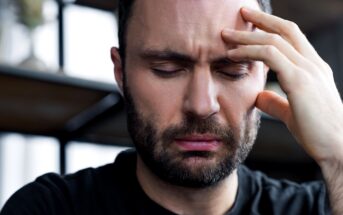 A man with a beard is closing his eyes, furrowing his brows, and touching his forehead with his hand, appearing to be in pain or deep thought. He is wearing a black shirt and is in an indoor setting with blurred shelves in the background.