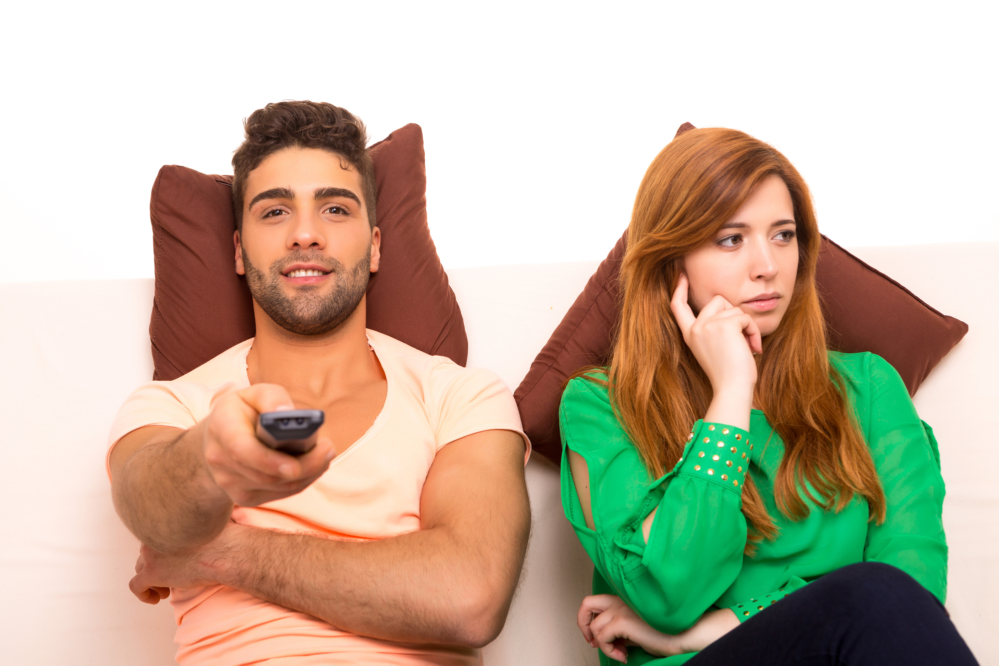 A man and a woman are seated on a couch, with pillows behind their backs. The man, wearing an orange shirt, is holding a TV remote and smiling. The woman, with long red hair in a green blouse, has her head resting on her hand and appears bored or disinterested.