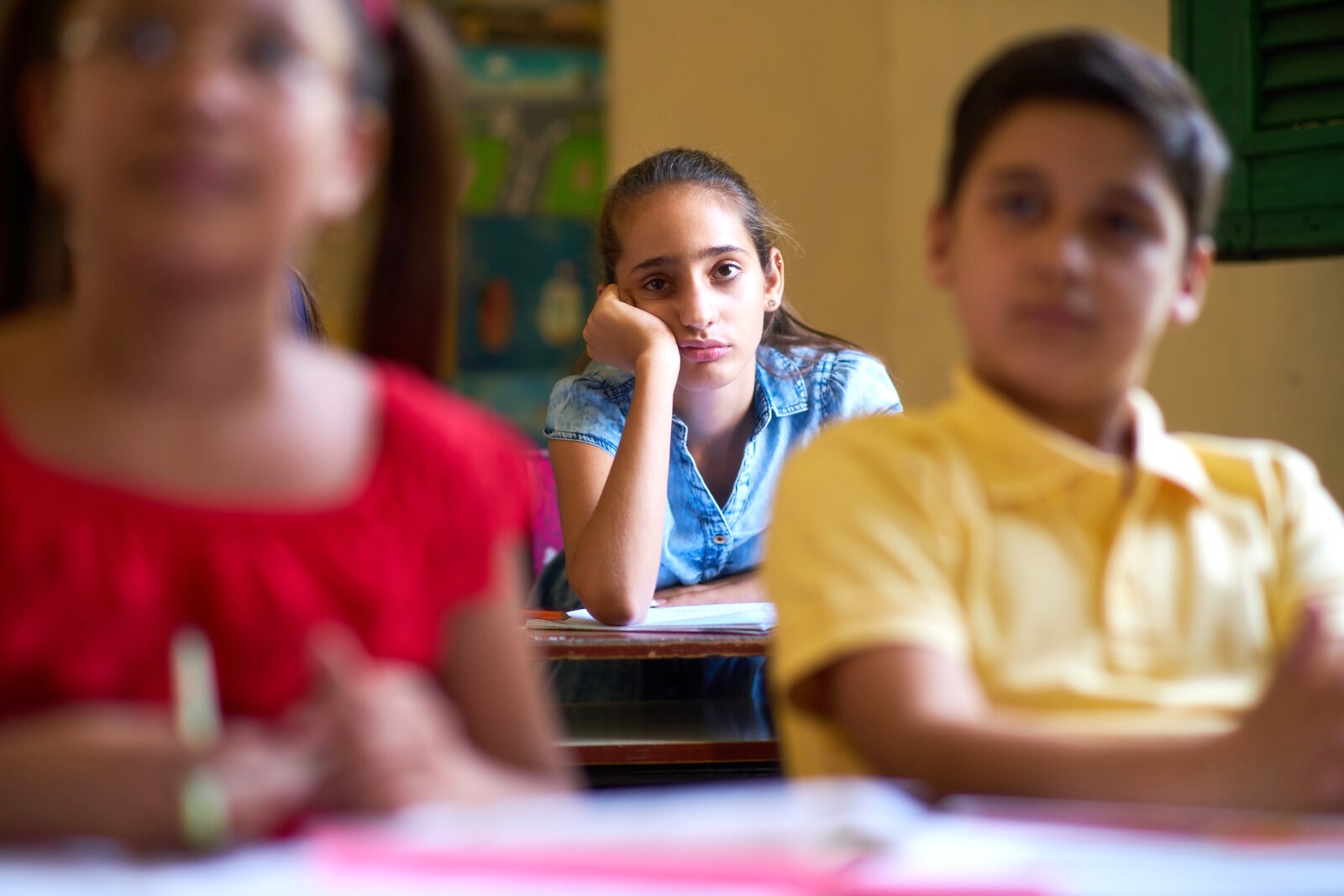 A girl in a blue shirt rests her head on her hand and looks thoughtfully ahead while sitting at a desk in a classroom. She is slightly blurred while two other students in the foreground, one in a red shirt and one in a yellow shirt, are in sharper focus.