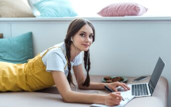 A young woman with braided hair lies on a bed, looking at her laptop while writing in a notebook. She is wearing earphones, a white t-shirt, and a yellow dress. Behind her, there are colorful pillows on a window seat. A plate of cookies and a smartphone are nearby.