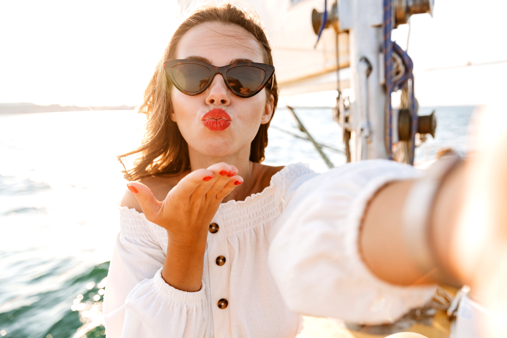 A person with long hair wearing sunglasses and a white off-shoulder top blows a kiss towards the camera. They are outdoors on a boat with sunlight in the background, creating a bright and cheerful atmosphere.