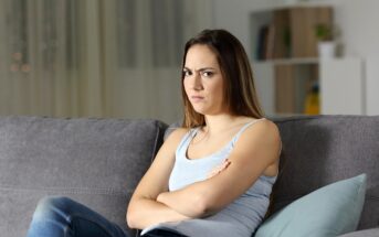 A young woman with long brown hair sits on a grey couch with her arms crossed and a frustrated expression on her face. She is wearing a light blue tank top and jeans. The background features a blurred bookshelf and curtains.
