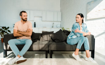 A man and a woman are sitting on opposite ends of a dark grey sofa in a bright, modern living room. Both appear to be upset, with the man crossing his arms and the woman turning away. The room features a white brick wall, cabinets, and minimal decor.