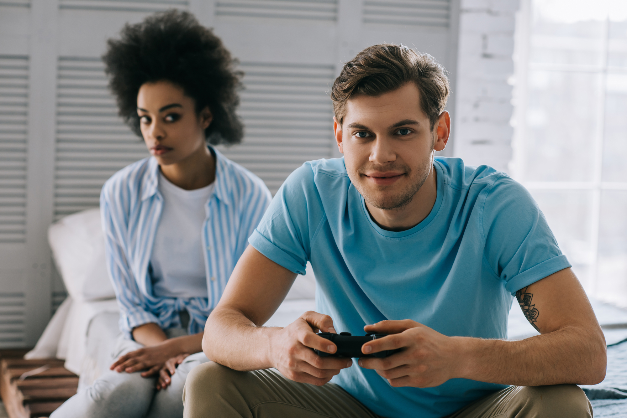 A man in a blue t-shirt plays a video game using a controller while sitting on a bed. Behind him, a woman with curly hair and in a striped shirt sits slightly turned to the side, looking away with a neutral expression. The room is bright with white decor.