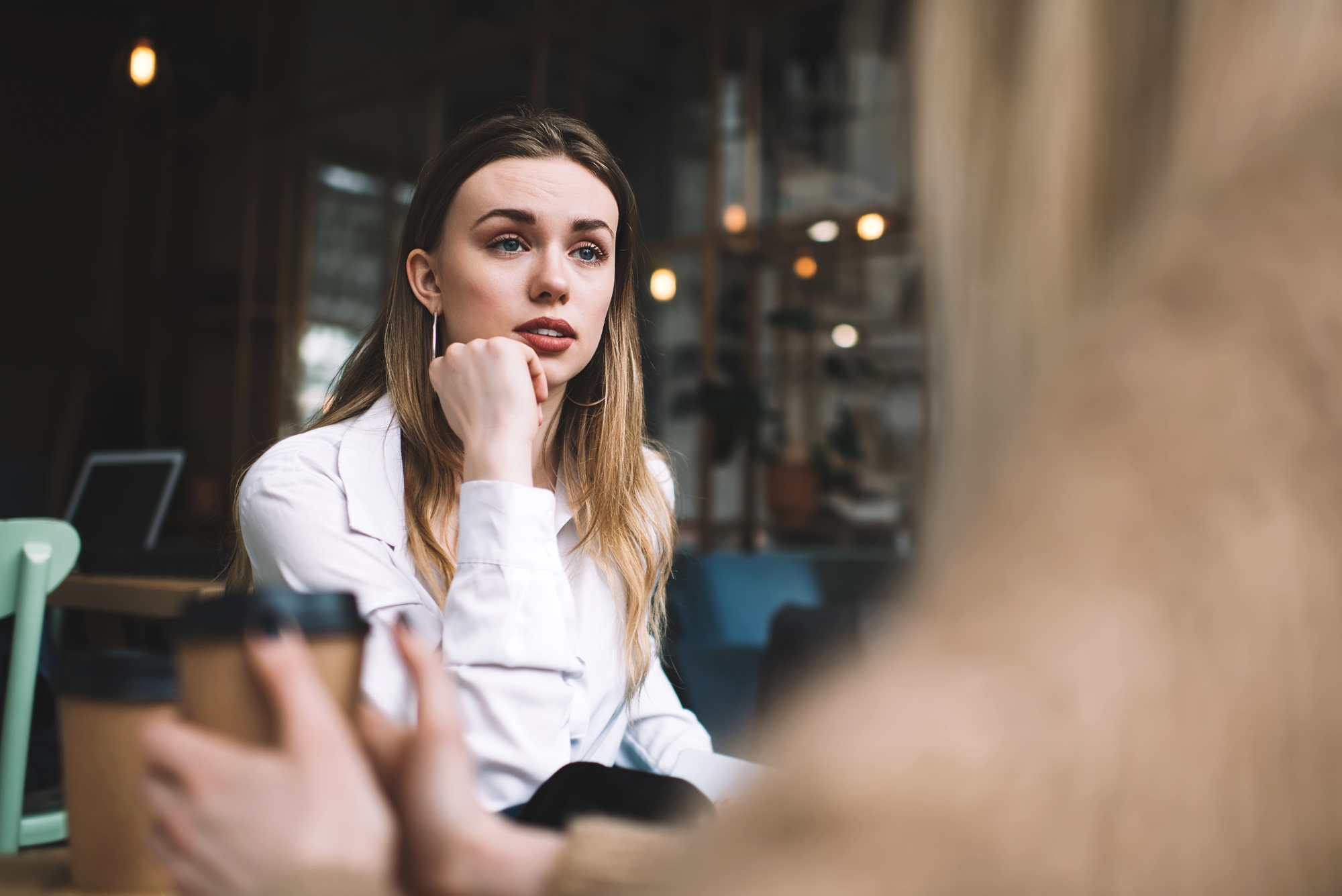 A young woman with long, blonde hair, wearing a white shirt, sits in a cafe with her hand resting on her chin, attentively listening to someone off-camera. She has a focused expression. The background shows a cozy, dimly lit cafe setting.