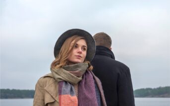 A woman in a gray hat, beige coat, and striped scarf looks contemplative, facing away from a man in a black coat. They stand outdoors with a cloudy sky and water in the background. The scene feels somber and introspective.