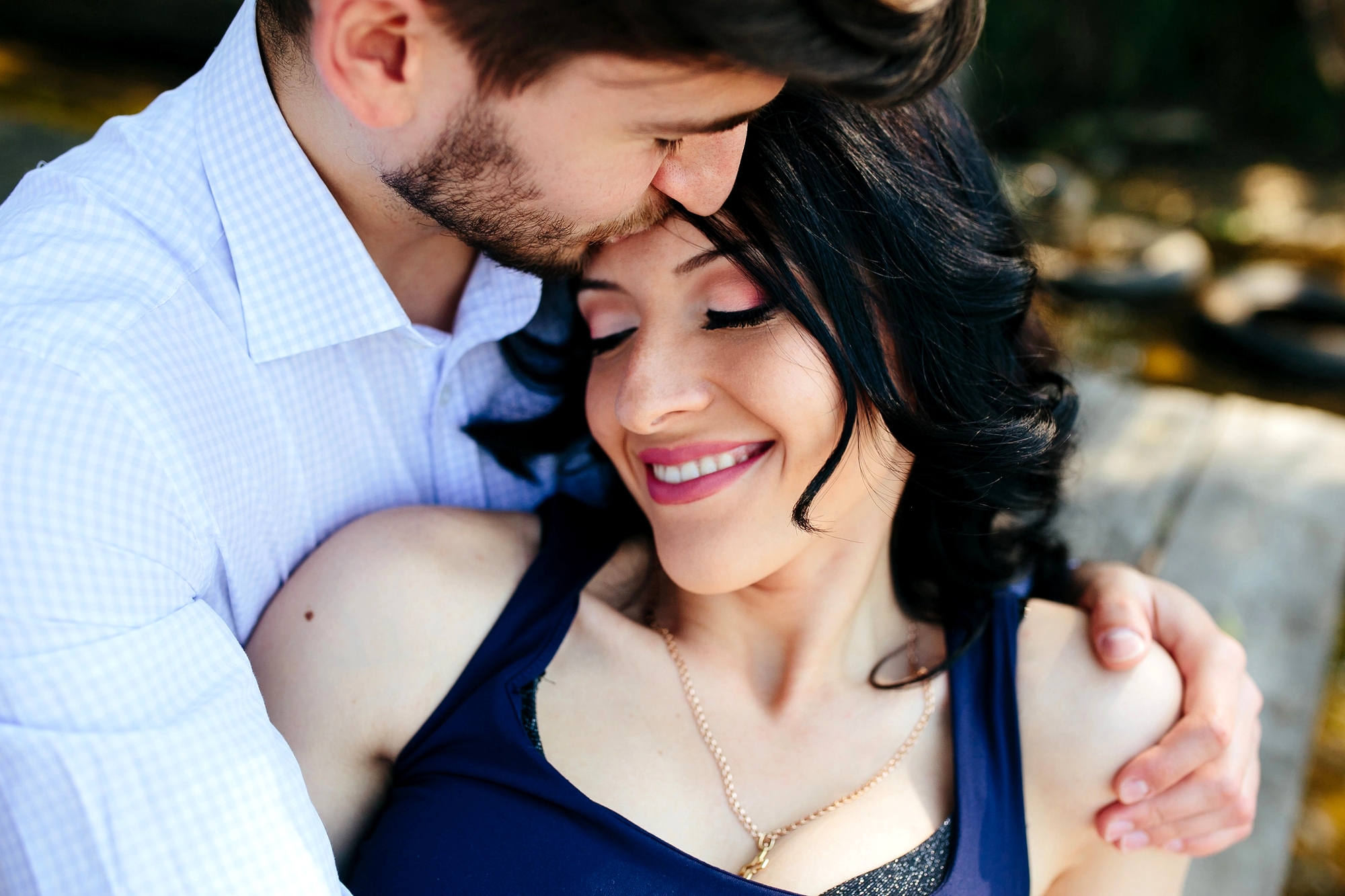 A man with short hair kisses the forehead of a woman with long, dark hair. The woman is smiling with her eyes closed, wearing a navy blue top and a necklace. The man is wearing a light blue checkered shirt. They seem to be outdoors, possibly enjoying a sunny day.