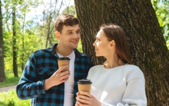A man and a woman stand outdoors by a tree, smiling at each other and holding coffee cups. The man wears a blue and black checkered shirt and the woman wears a white sweatshirt. The background features lush greenery and a sunny sky.