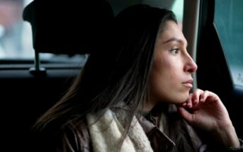 A woman with long dark hair sits in a car, resting her chin on her hand, and gazes out the window contemplatively. She is wearing a brown jacket with a light-colored scarf. The interior of the car is dimly lit.