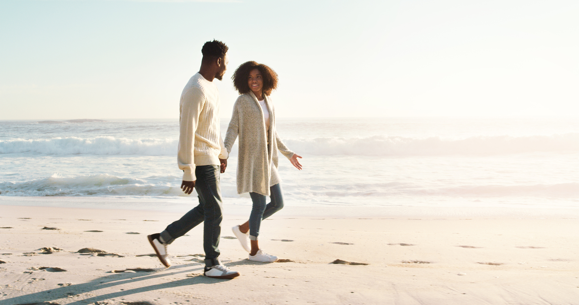 A couple walks hand in hand along a sandy beach with waves gently crashing in the background. Both are wearing light sweaters and jeans, and they appear to be engaged in a pleasant conversation under the soft sunlight.