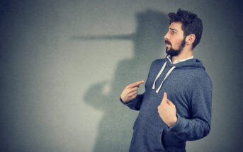 A bearded man in a blue hoodie stands against a gray background, pointing to himself with both thumbs. A shadow on the wall behind him has an exaggerated long nose, evoking the imagery of Pinocchio, suggesting dishonesty or lying.