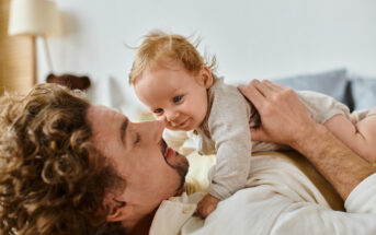 A person with curly hair lies on their back, holding a baby with light-colored hair on their chest. They gaze lovingly at each other, with the baby smiling and dressed in light-colored clothing. The background is a cozy indoor setting with soft lighting.