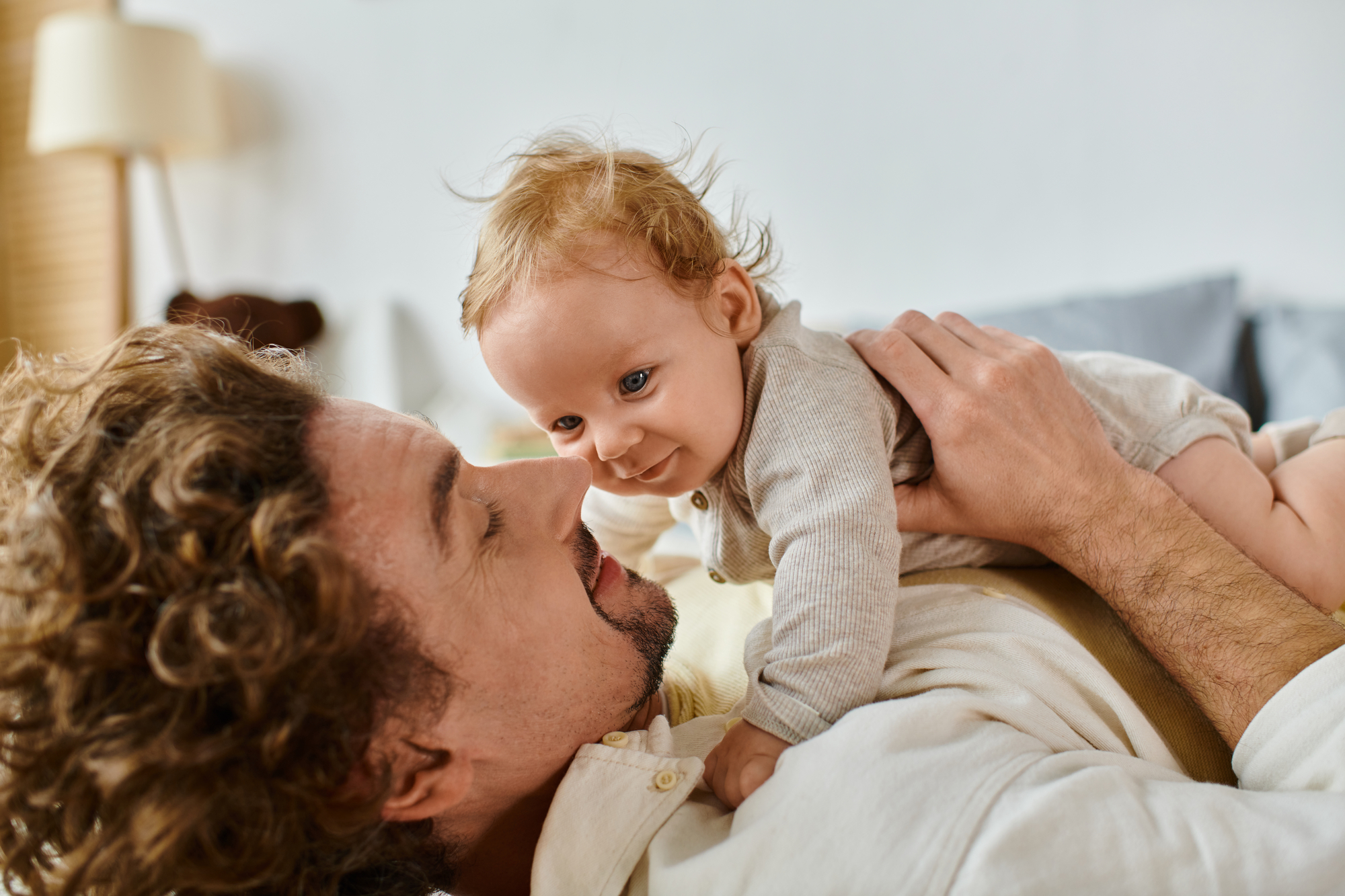 A person with curly hair lies on their back, holding a baby with light-colored hair on their chest. They gaze lovingly at each other, with the baby smiling and dressed in light-colored clothing. The background is a cozy indoor setting with soft lighting.