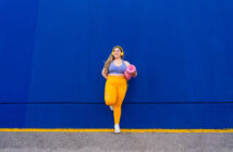 A woman stands against a bright blue wall, looking off to the side. She is wearing a yellow headband, workout top, and leggings, and is holding a pink yoga mat. The ground has a yellow line running parallel to the wall.