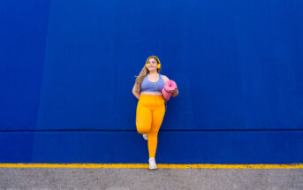 A woman stands against a bright blue wall, looking off to the side. She is wearing a yellow headband, workout top, and leggings, and is holding a pink yoga mat. The ground has a yellow line running parallel to the wall.