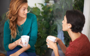 Two women are sitting and holding white mugs while engaging in conversation. The woman on the left has long, wavy hair and wears a teal top, while the woman on the right has short hair and wears a reddish-brown top. Lush green plants are visible in the background.