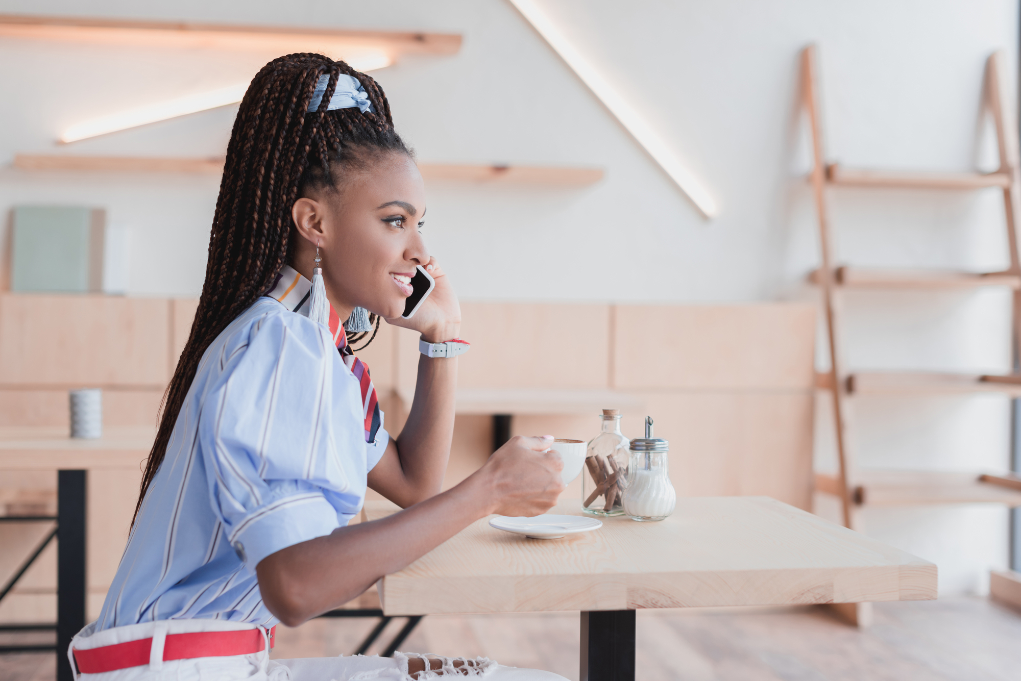 A woman with long braided hair sits at a wooden table in a modern café. She is holding a cup of coffee in one hand and talking on a mobile phone with the other. She is dressed in a light blue shirt with white stripes, and the background features minimalist decor.