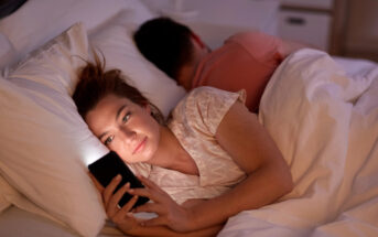 A woman is lying in bed at night, using her smartphone which is illuminating her face. She appears focused on the screen. Beside her, another person is lying down, facing away and seemingly asleep. They are under white bedcovers.