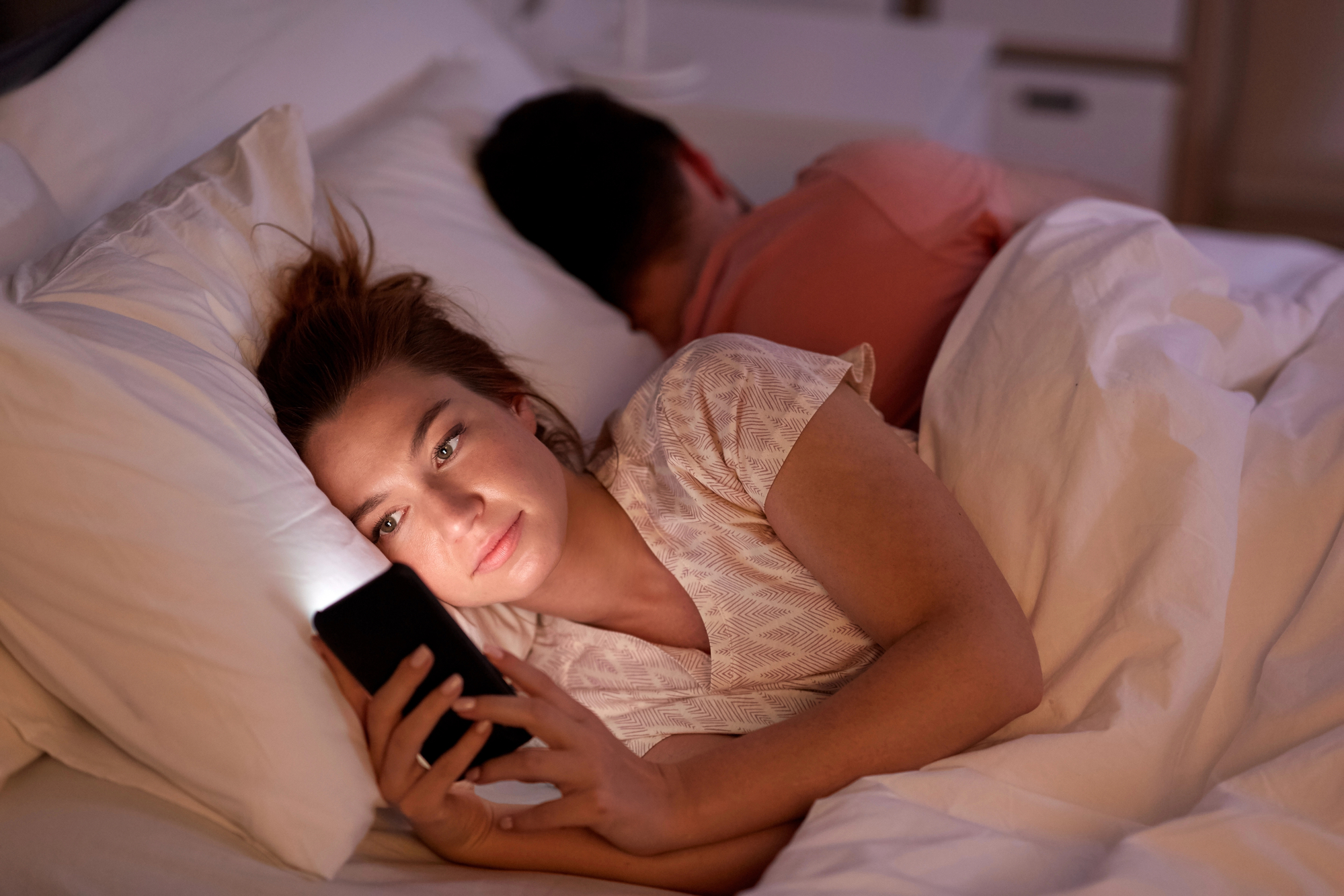 A woman is lying in bed at night, using her smartphone which is illuminating her face. She appears focused on the screen. Beside her, another person is lying down, facing away and seemingly asleep. They are under white bedcovers.