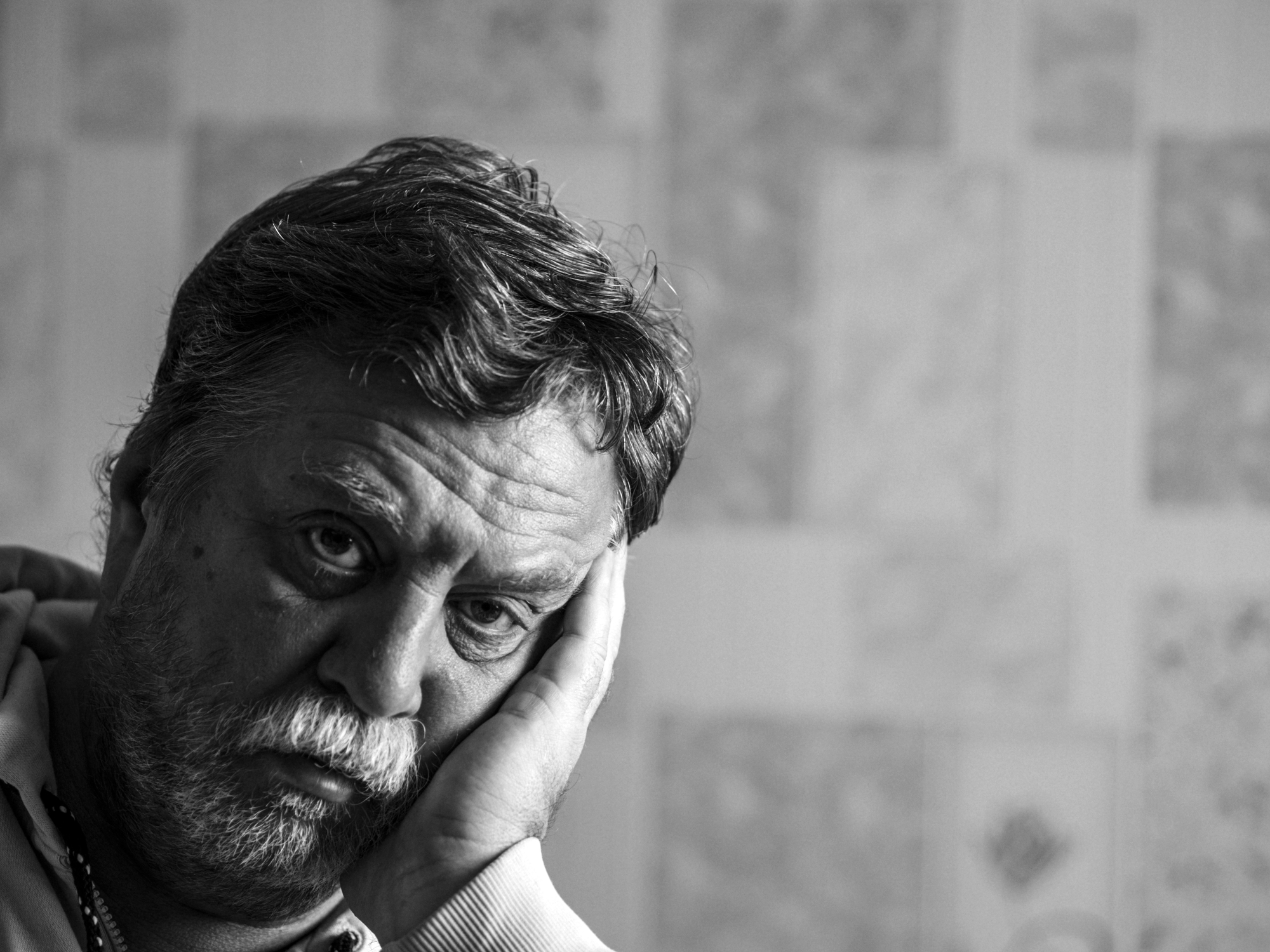 A grayscale image of an older man resting his head on his hand with a pensive expression. His face is framed by tousled hair and a beard. The background is softly blurred, featuring a pattern of various shades.