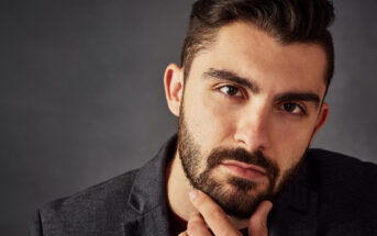 Close-up portrait of a man with a well-groomed beard and dark hair, wearing a dark blazer. He is looking directly at the camera with a thoughtful expression, his hand resting on his chin against a dark, neutral background.