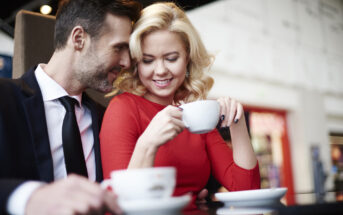 A couple is sitting closely together at a table in a cafe. The man, wearing a black suit and tie, is leaning in and smiling, while the woman, in a red dress with blonde hair, is holding a cup of coffee and smiling down at it. The background is softly blurred.