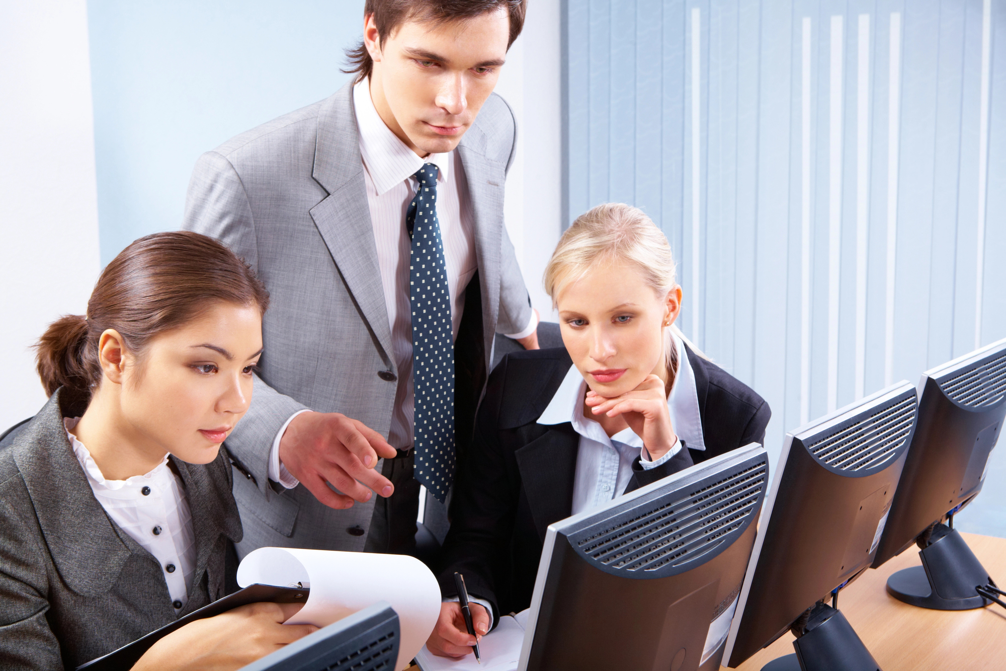 Three business professionals are focused on computer monitors. A man in a gray suit gestures towards the screens, while two women, also in business attire, closely observe. The group appears engaged in a serious discussion or analysis. Office blinds are visible in the background.