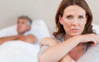 A woman with shoulder-length brown hair sits with her arms crossed and an expression of concern or sadness. In the background, a man with gray hair lies on a bed, facing partially away from her. The setting appears to be a bedroom.